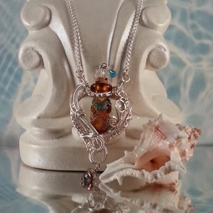 Op de afbeelding: Een zilveren ketting met een hartvormige hanger met een bruine glazen fles met blauwe en gouden accenten. De hanger is gemaakt van draad dat om de fles is gewikkeld. De ketting wordt getoond op een wit voetstuk met een schelp.