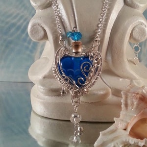 Puede incluir: Un collar de cadena plateada con un colgante de botella de vidrio azul en forma de corazón. La botella está envuelta en alambre plateado y tiene una cadena plateada con cuentas transparentes que cuelgan de ella.