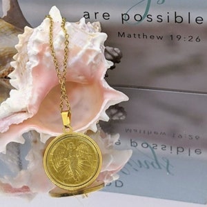 Puede incluir: Collar de oro con un colgante redondo con un dise&ntilde;o de &aacute;ngel. El colgante est&aacute; sobre una concha con el texto "With God all things are possible Matthew 19:26" en el fondo.