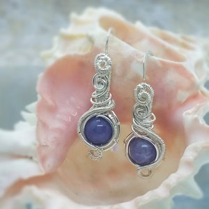 Puede incluir: Un par de pendientes de alambre plateado con cabujones de piedras preciosas púrpuras. Los pendientes cuelgan de una concha.