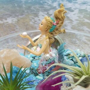 Può includere: Una statuetta di sirena in miniatura con capelli biondi e coda turchese si trova in un terrario di vetro con sabbia blu, pietre bianche, una fetta di agata rosa e una pianta aerea verde.