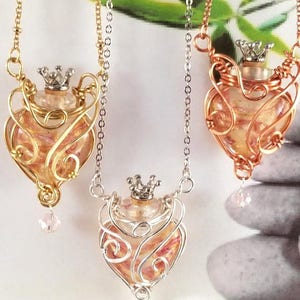 Puede incluir: Tres botellas de vidrio en forma de corazón con una corona en la parte superior, cada una encerrada en una jaula de alambre. Las botellas están llenas de purpurina rosa y blanca y cuelgan de cadenas plateadas, doradas y de cobre. Las botellas cuelgan sobre un fondo de hojas verdes y piedras grises.