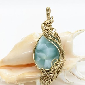 Puede incluir: Un colgante de alambre dorado con una piedra preciosa azul claro y blanca. El colgante se muestra en una concha.