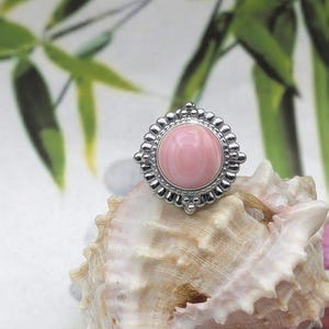 May include: A silver ring featuring a large, round, pink gemstone. The ring rests on a seashell, against a blurred green and white backdrop. The gemstone is encircled by a decorative silver setting.