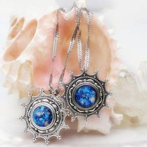 May include: Two silver-toned pendant necklaces with a sunburst design. Each pendant features a round, blue, speckled center. The necklaces are displayed on a light pink seashell, creating a contrast of colors and textures. The chains are delicate.
