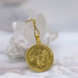 Puede incluir: Collar de cadena dorada con un colgante redondo que representa un &aacute;ngel con alas y un halo. El colgante tiene un dise&ntilde;o detallado y un acabado brillante.