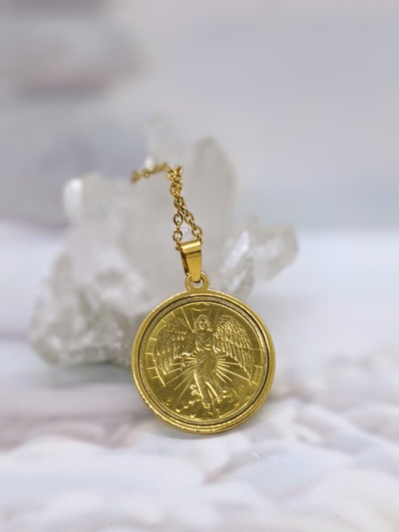 Puede incluir: Collar de cadena dorada con un colgante redondo que representa un &aacute;ngel con alas y un halo. El colgante tiene un dise&ntilde;o detallado y un acabado brillante.