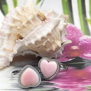 May include: Two silver rings, each featuring a pink heart-shaped stone. The rings are displayed with a seashell and a pink and white floral design in the background. A romantic and feminine jewellery item.