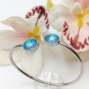 May include: A silver bangle bracelet with two light blue crystal accents. The bracelet is on a white background with white flowers.