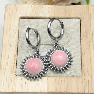 May include: A pair of silver hoop earrings with a pink, circular center stone. The stone is surrounded by a silver, sunburst-like design. The earrings are displayed in a wooden box.