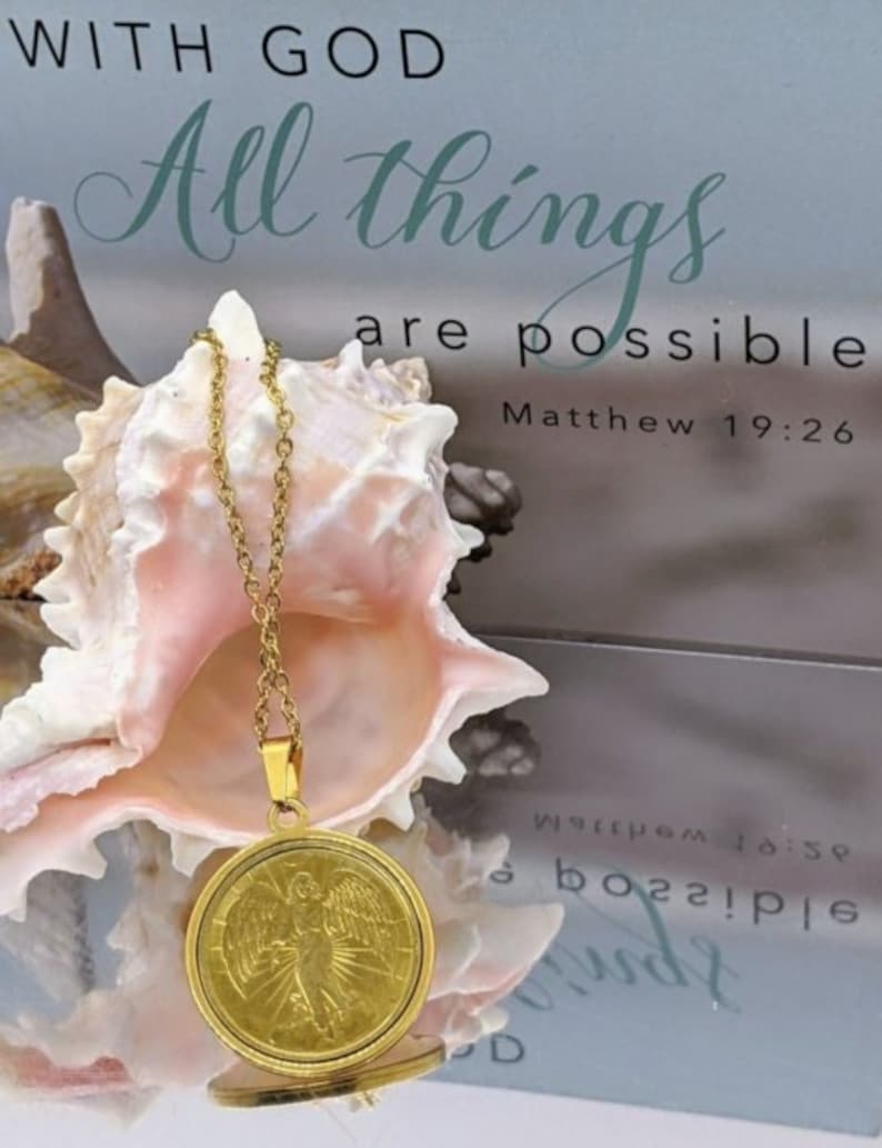 Puede incluir: Collar de oro con un colgante redondo con un dise&ntilde;o de &aacute;ngel. El colgante est&aacute; sobre una concha con el texto "With God all things are possible Matthew 19:26" en el fondo.