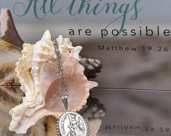 May include: A silver necklace with a silver oval pendant featuring a guardian angel design. The pendant has the words "Guardian Angel" inscribed on it. The necklace is resting on a seashell. The background is a card with the text "With God All things are possible Matthew 19:26".