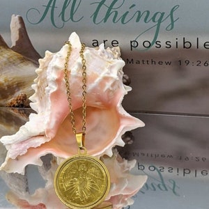 Puede incluir: Collar de oro con un colgante redondo que presenta un &aacute;ngel con alas. El colgante cuelga de una concha de mar sobre un fondo blanco con el texto "Con Dios todo es posible Mateo 19:26".