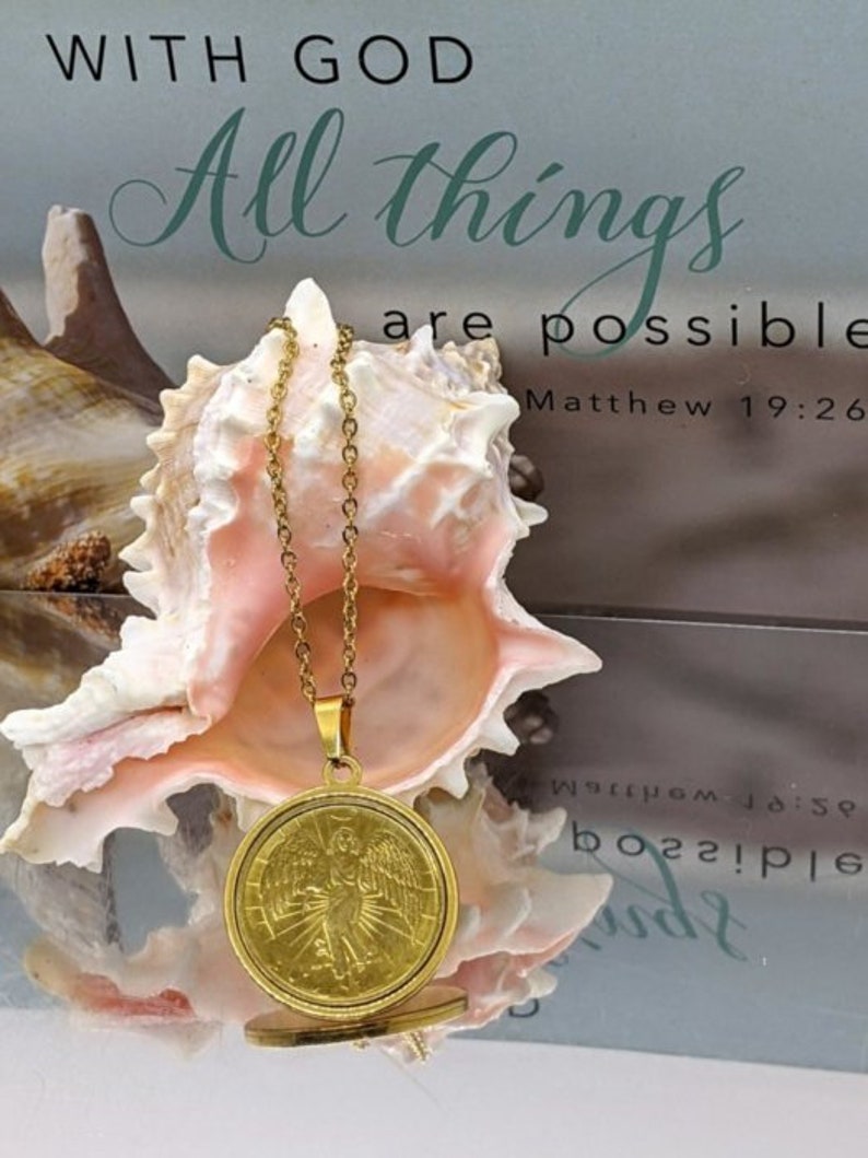 Puede incluir: Collar de oro con un colgante redondo que presenta un &aacute;ngel con alas. El colgante cuelga de una concha de mar sobre un fondo blanco con el texto "Con Dios todo es posible Mateo 19:26".