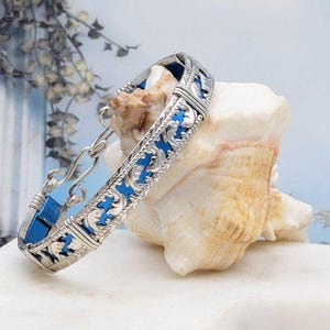 May include: A silver bracelet with a blue enamel inlay and a decorative design. The bracelet is on a white surface with a seashell.