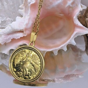 Puede incluir: Collar de cadena dorada con un colgante redondo que presenta un &aacute;ngel con alas. El colgante tiene un dise&ntilde;o detallado y est&aacute; colocado sobre un fondo de concha rosa.