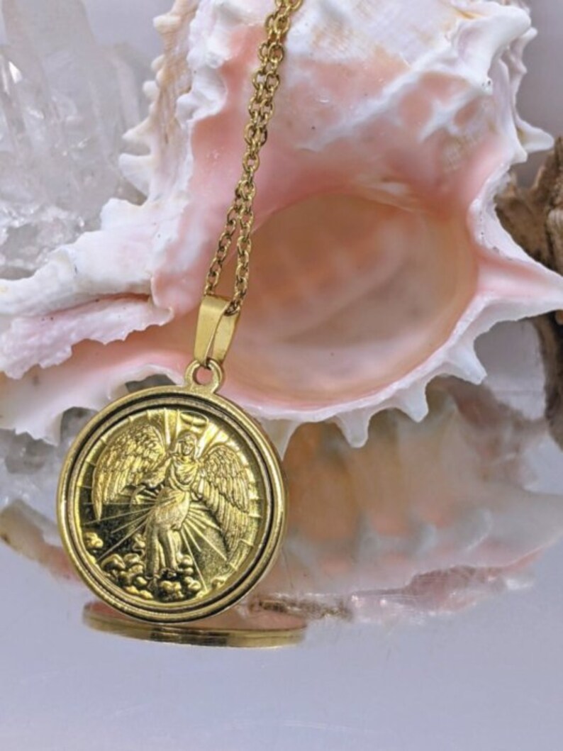 Puede incluir: Collar de cadena dorada con un colgante redondo que presenta un &aacute;ngel con alas. El colgante tiene un dise&ntilde;o detallado y est&aacute; colocado sobre un fondo de concha rosa.