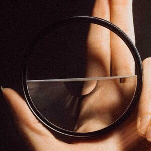 May include: A circular black camera filter with a thin, clear glass element held in a hand against a dark background. A beam of light refracts through the glass, creating a rainbow effect. The filter is designed for photography.