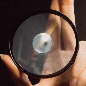 May include: A circular camera lens filter with a black frame is held between fingers against a dark background. The filter's glass is clear, with a central, reflective element. The image is well-lit, highlighting the filter's design.