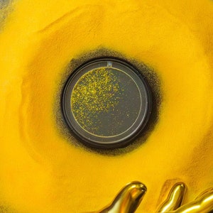 Pode incluir: Uma mão dourada estendendo-se para um círculo preto com glitter dourado dentro de um círculo amarelo sobre um fundo preto.