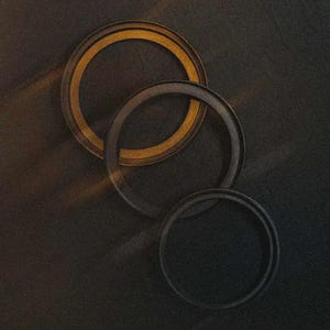 Puede incluir: Tres marcos circulares superpuestos en diferentes tonos de negro y dorado están dispuestos diagonalmente sobre un fondo oscuro. El marco superior tiene un anillo interior dorado, mientras que el marco central es gris oscuro. El marco inferior es negro.