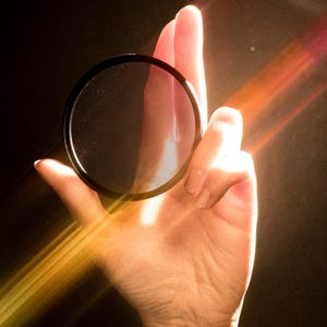 Peut inclure: Un filtre d'objectif d'appareil photo noir et circulaire tenu dans une main sur fond sombre. Le filtre est transparent avec un bord noir. Des rayons de lumière traversent l'image, créant une lueur dorée et chaleureuse.