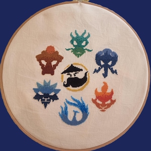 May include: A cross-stitch embroidery hoop displaying seven colourful, pixelated symbols. The symbols include a dragon, a ninja with a moustache, a phoenix, and other abstract shapes in shades of blue, orange, and green. The embroidery is set against a cream-coloured fabric.