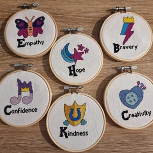 May include: Six embroidery hoops with different designs and words. The designs include a butterfly, a crescent moon and star, a musical note, a shield with a horseshoe, a lightning bolt, and a heart. The words are "Empathy", "Hope", "Confidence", "Kindness", "Bravery", and "Creativity".