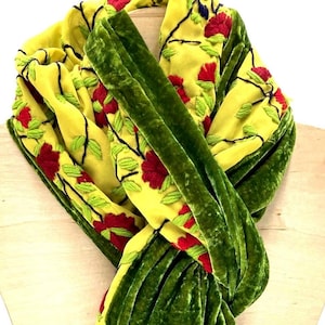 May include: A bright yellow scarf featuring embroidered red flowers and green leaves on black stems. The scarf is accented with wide, textured dark green velvet borders that run along the length of the fabric, creating a layered appearance.