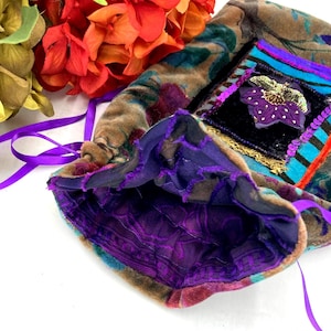 May include: A velvet drawstring pouch featuring a multi-colored floral pattern in shades of brown, blue, and purple. The bag includes a purple satin ribbon closure, a black velvet square patch with a purple flower appliqué, and a purple lining.