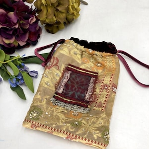 May include: A drawstring pouch made of gold-toned fabric with an intricate floral pattern, embellished with silver sequins and beads. It features a central rectangular patch in deep burgundy and dark teal, finished with a dark velvet drawstring closure.