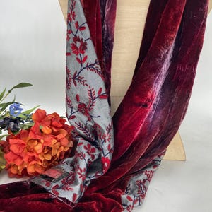 May include: A long scarf featuring deep red velvet fabric on one side and a light gray fabric on the other, decorated with an embroidered pattern of red flowers and dark blue stems. The scarf is draped over a light-colored display form.