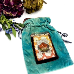 May include: A turquoise velvet drawstring pouch featuring a rectangular patch on the front. The patch displays a white and orange geometric pattern, a central orange floral motif, and is framed by dark brown velvet trim and gold lace.