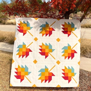 May include: A white quilt with a patchwork design of colorful maple leaves. The leaves are made up of squares in shades of red, orange, yellow, and blue. The quilt is hanging on a clothesline in front of a tree with red leaves.