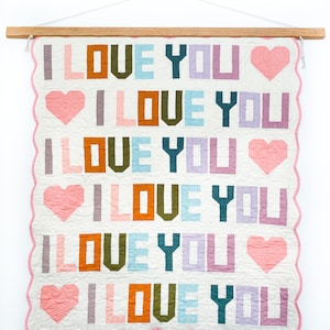 May include: A quilted wall hanging with the words "I LOVE YOU" in colorful block letters and heart shapes. The quilt has a scalloped pink border and is suspended from a wooden rod.