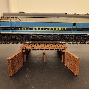 May include: A model train locomotive in blue and black, with the words "BALTIMORE AND OHIO" printed on the side, sits on a track above a brown wooden bridge. The train is a detailed replica of a real-life train.