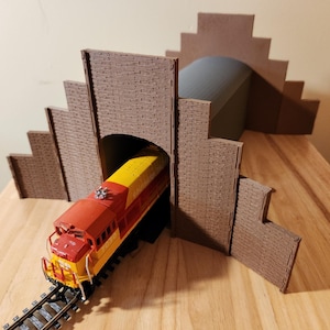 May include: A model train locomotive, red and yellow, emerges from a brown tunnel. The tunnel is part of a larger brown structure with a brick-like texture, set on a wooden surface. The train is on a black track.