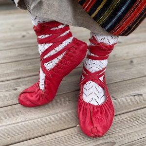 May include: A pair of red leather shoes with crisscross straps, paired with white lace socks. The shoes have a rounded toe and are gathered at the top. The socks are visible above the shoes, creating a layered look.