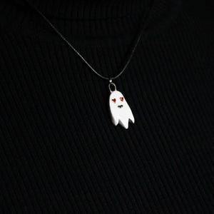 Peut inclure: Un pendentif fantôme blanc avec des yeux et une bouche rouges est suspendu à une chaîne argentée. Le pendentif fantôme est le point central sur un fond noir. Le collier est un accessoire simple mais effrayant.