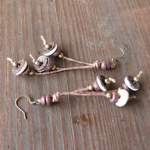 May include: Handmade earrings featuring a combination of brown, white, and brass-colored beads. The earrings have a rustic, bohemian style, with a natural cord connecting the beads and a simple hook closure. The design incorporates button-like elements.