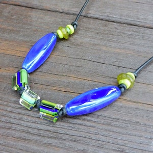 May include: A handmade necklace featuring vibrant blue, green, and clear glass beads. The necklace has elongated blue beads and square, multi-colored beads, strung on a black cord. Small, round green beads accent the design, creating a unique accessory.