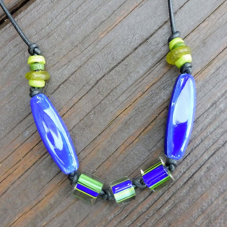 May include: A handmade necklace featuring a black cord strung with blue, green, and clear glass beads. The necklace has two large, oblong blue beads and several smaller, square beads with green and blue stripes. The necklace is displayed on a wooden surface.