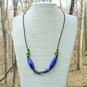May include: A handmade necklace featuring a black cord with blue, green, and black beads. The necklace has two large, oval-shaped blue beads and several smaller green and black beads. The necklace is displayed on a mannequin.