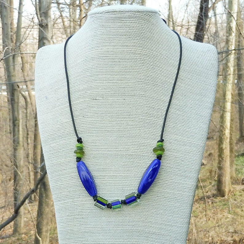 May include: A handmade necklace featuring a black cord with blue, green, and black beads. The necklace has two large, oval-shaped blue beads and several smaller green and black beads. The necklace is displayed on a mannequin.
