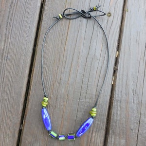 May include: A handmade necklace featuring a black cord and a series of blue and green glass beads. The necklace has a simple design with elongated blue beads and smaller, square green beads. The cord is tied at the top.