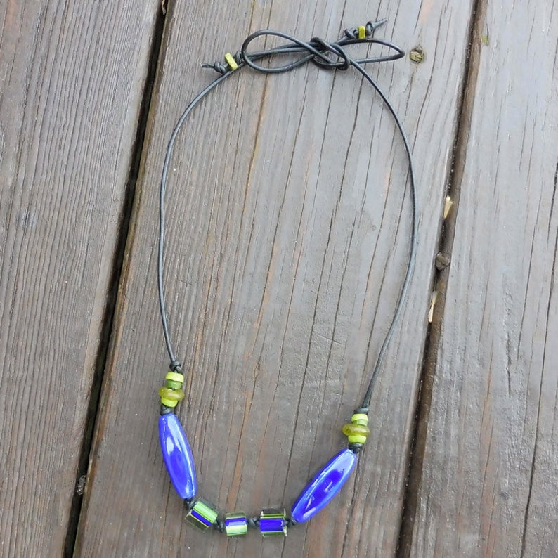 May include: A handmade necklace featuring a black cord and a series of blue and green glass beads. The necklace has a simple design with elongated blue beads and smaller, square green beads. The cord is tied at the top.