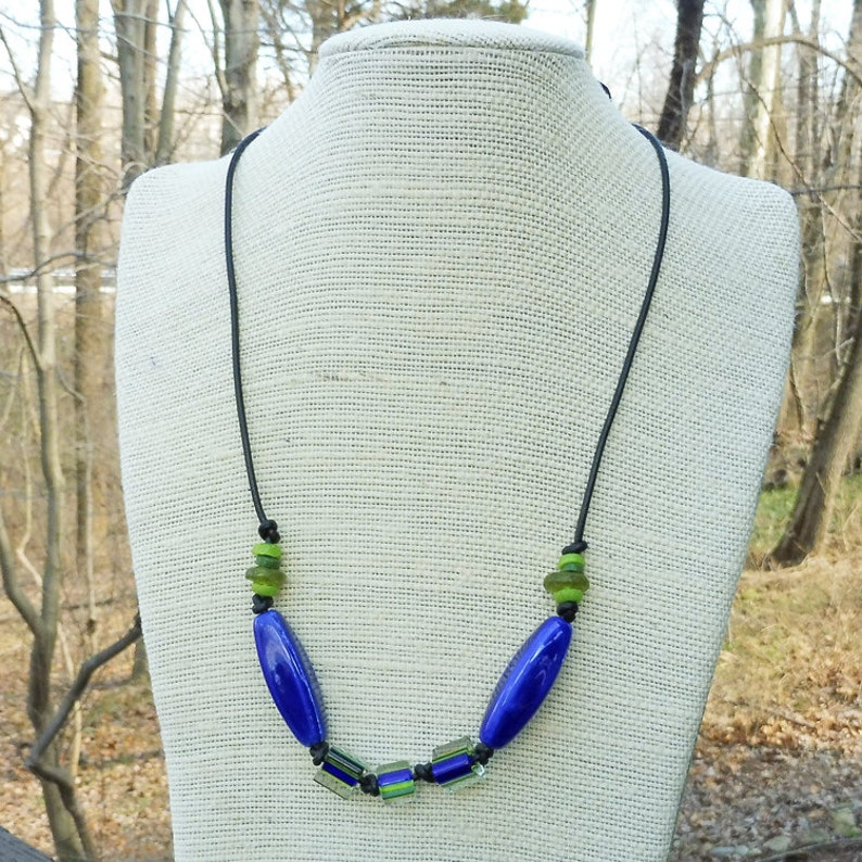 May include: A handcrafted necklace featuring a black cord with blue and green glass beads. The necklace has two large, oval blue beads and smaller green and blue rectangular beads. The beads are strung on a black cord.