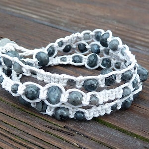 May include: A stack of three handmade bracelets. Each bracelet features gray, round beads strung on a white, woven cord. The bracelets are displayed on a wooden surface, showcasing the natural tones of the materials.