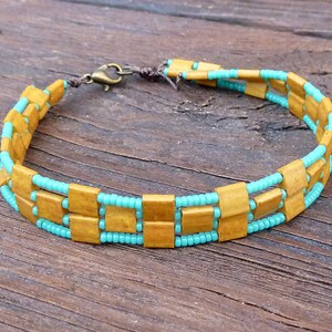 May include: A handmade bracelet featuring rectangular wooden beads in a warm, golden-brown hue, interspersed with rows of small, turquoise seed beads. The bracelet is secured with a bronze-colored clasp and brown cord.