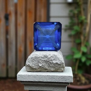 May include: A large, faceted, square-cut, deep blue gemstone sits atop a rough-hewn stone, which is placed on a gray, textured pedestal. The gemstone's facets reflect light, creating a brilliant display. The background features a wooden fence and greenery.